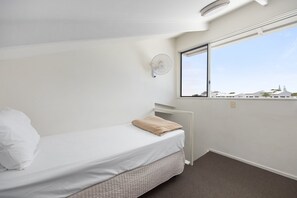 Un letto singolo con materasso bianco e coperta beige, una ventola a muro e una finestra con vista sugli edifici.