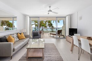 A modern living room with a sofa, coffee table, and TV, leading to a balcony with a view of the sea.