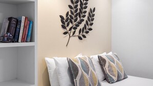 Una habitación con una cama blanca, almohadones estampados, una estantería con libros y un adorno en forma de hoja en la pared.