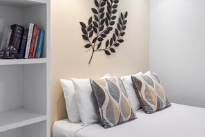 A room with a white bed, patterned pillows, a bookshelf with books, and a wall-mounted leaf decoration.