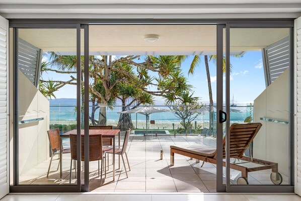 A glass-enclosed balcony with a view of the beach, a table, and two chairs.