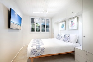 A bedroom with a bed, a TV mounted on the wall, a window with shutters, and a ceiling fan.
