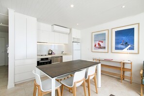 Cozinha moderna com armários brancos, forno embutido e geladeira. Há uma área de jantar com mesa escura e cadeiras brancas. Duas fotos emolduradas estão na parede.