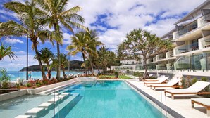 A swimming pool with lounge chairs, palm trees, and a modern building with balconies.