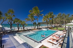 A pool area with a glass barrier, lounge chairs, and a view of the beach.