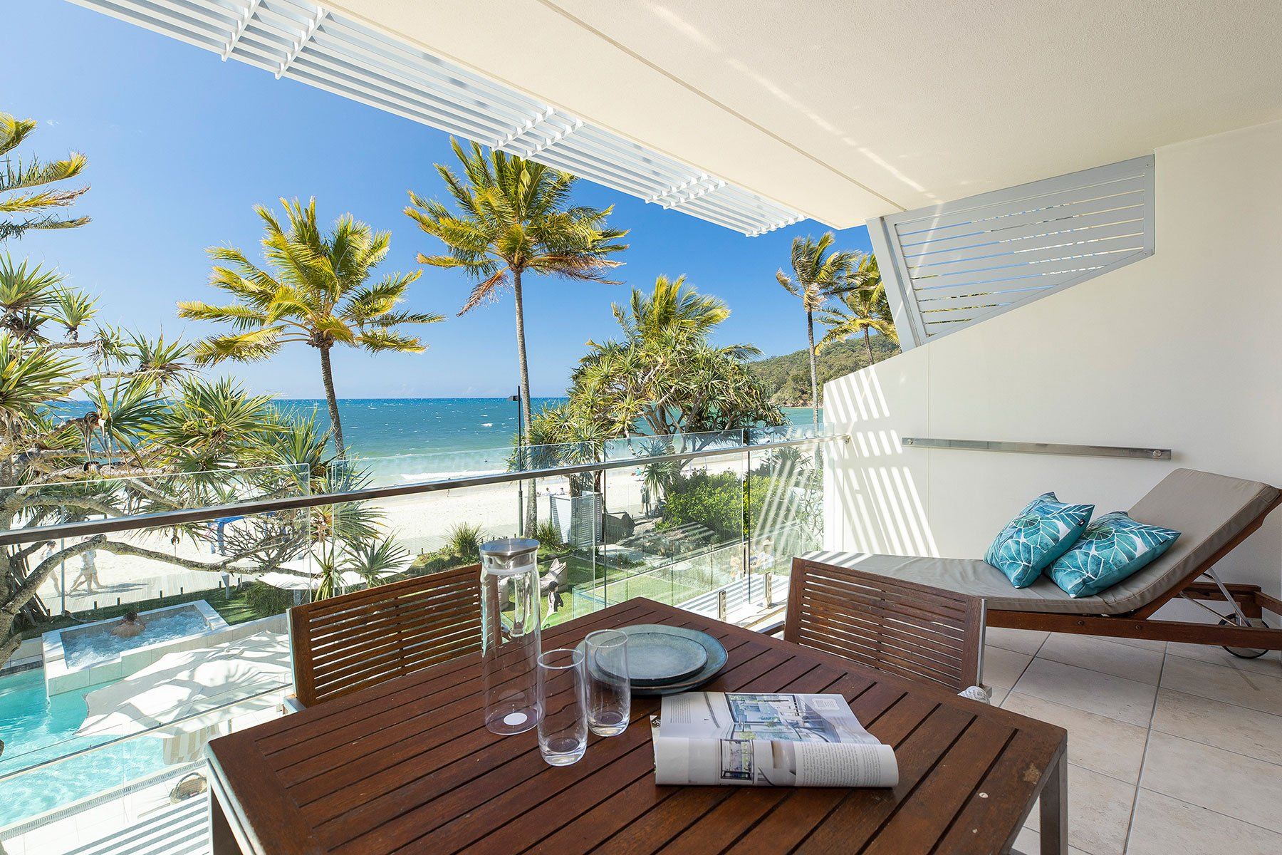 A balcony with a view of the beach and ocean, wooden furniture, and a glass railing.