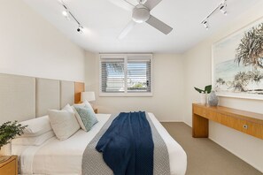 Une chambre à coucher comprenant un lit, une table de chevet, une commode, un ventilateur de plafond et une fenêtre avec des stores.