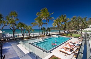 A pool area with a glass barrier, lounge chairs, and a view of the beach.
