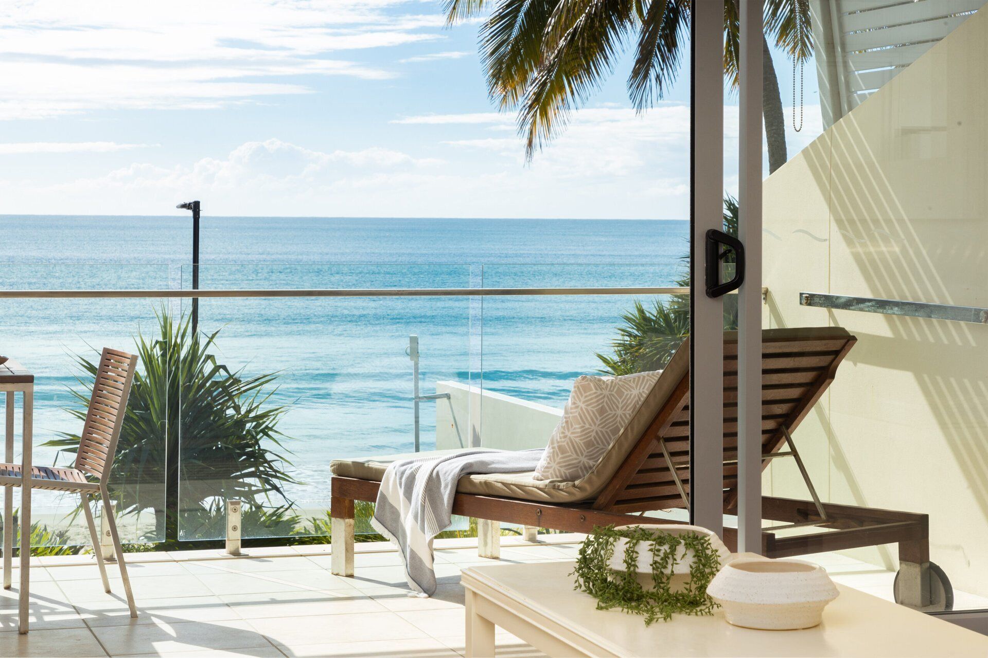 A balcony with a view of the ocean, a wooden lounge chair, a small table, and a potted plant.