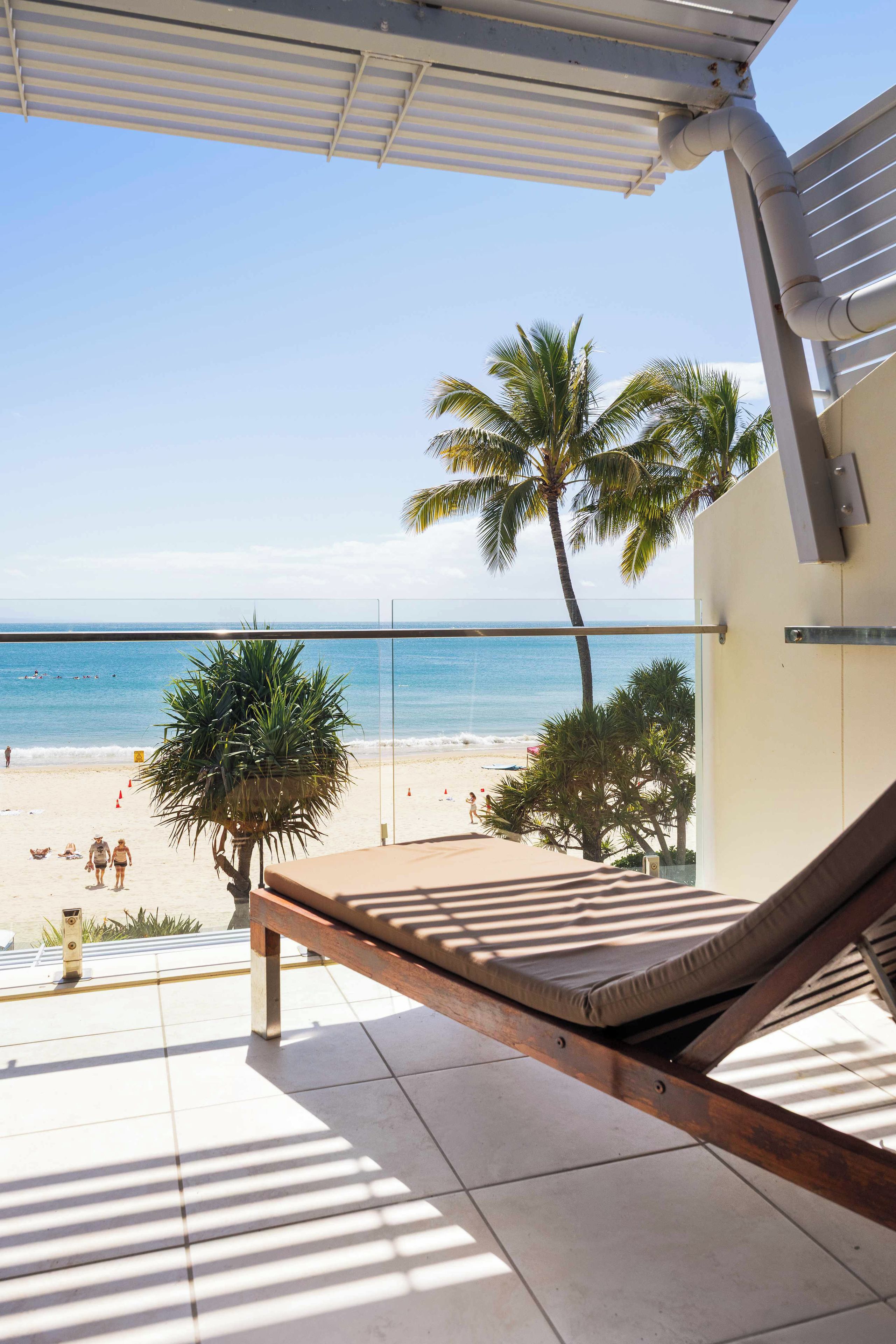 A beach view from a balcony with a lounge chair, palm trees, and a clear sky.