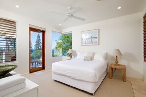 A bedroom with a bed, a ceiling fan, a window with blinds, a picture on the wall, and a small table with a lamp. - Hale Lani 2 - Oceanview Penthouse Paradise (Sunshine Beach)