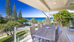 A wooden table with chairs and a glass partition, overlooking a coastal view with trees and a clear sky. - Hale Lani 2 - Oceanview Penthouse Paradise (Sunshine Beach)