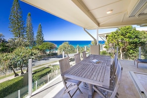 A wooden table with chairs and a glass partition, overlooking a coastal view with trees and a clear sky.