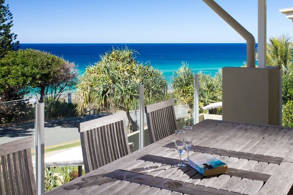 A wooden table with two glasses and a book, overlooking a coastal view with the ocean and greenery.