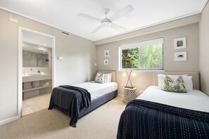 A bedroom with two beds, a ceiling fan, a window with a view of greenery, and a bathroom with a shower and sink.