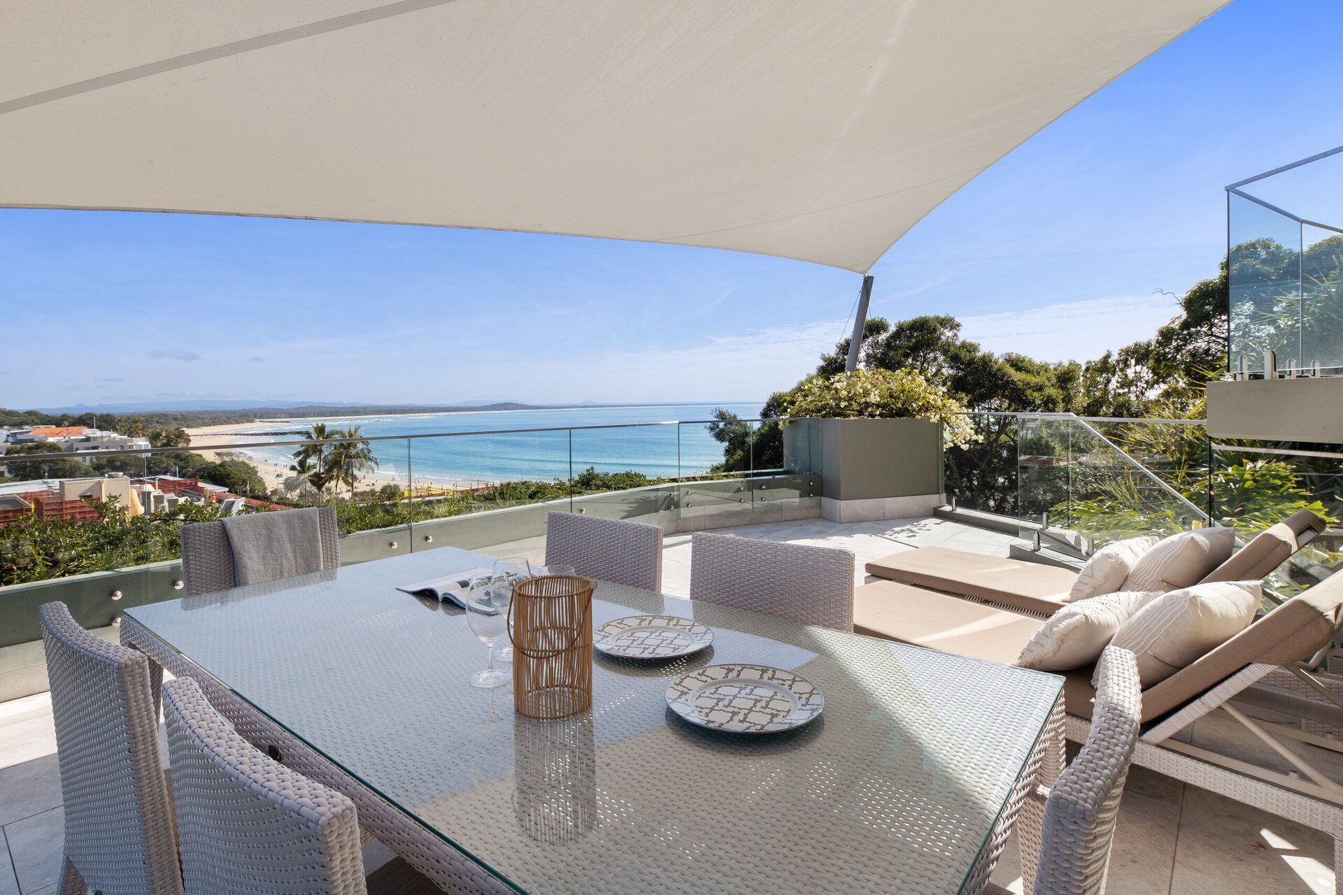 A glass-top table with wicker chairs, a glass balustrade, and a view of the beach and ocean.