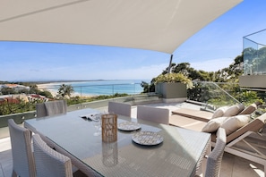 A glass-top table with wicker chairs, a glass balustrade, and a view of the beach and ocean.