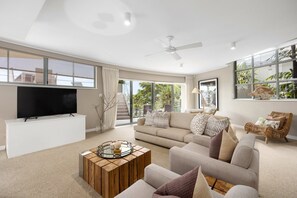 A modern living room with a flat-screen TV, a ceiling fan, and a large window offering a view of the outdoors.