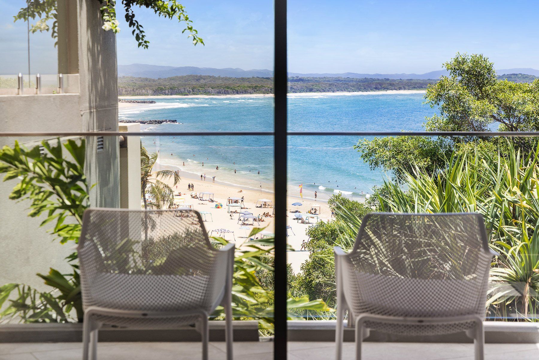 A balcony with two wicker chairs overlooking a beach with people and umbrellas.