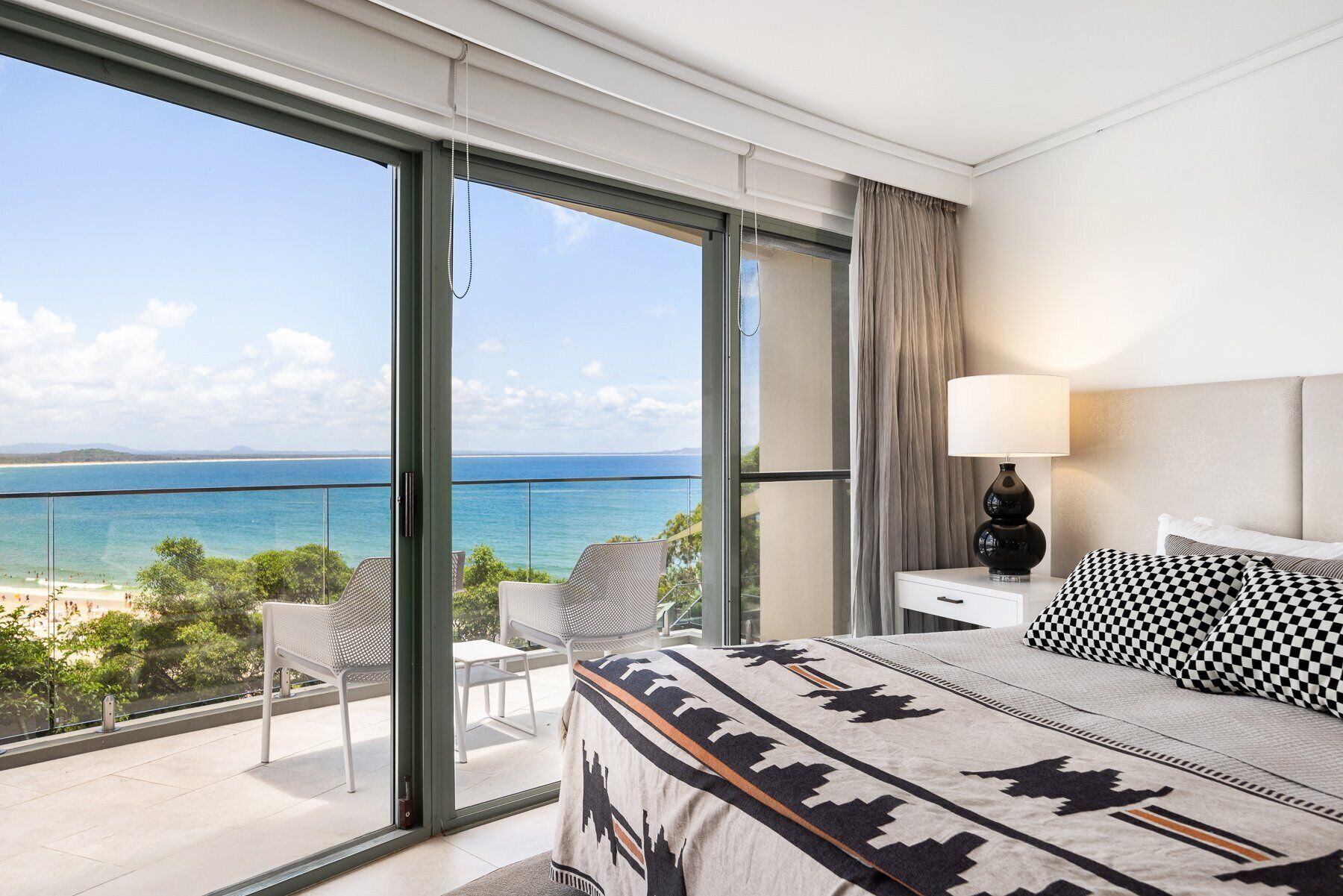 A hotel room with a large balcony, a bed with patterned bedding, a nightstand with a lamp, and a view of the sea.