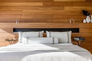 A modern bedroom with a wooden headboard, a bed with white bedding, and a small bedside table with a book.