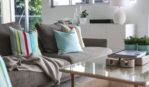 Ein modernes Wohnzimmer mit einem grauen Sofa, bunten Kissen, einem Glastisch und einer weißen Lampe.