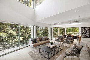 A spacious living area with a high ceiling, large windows, a sofa, chairs, a coffee table, and a dining table.