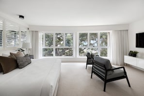 A modern bedroom with a large bed, a seating area, and multiple windows offering a view of trees.