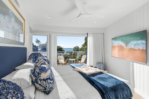 A bedroom with a bed, a ceiling fan, a painting, and a view of the sea.