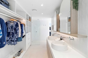 A modern bathroom with two sinks, a large mirror, and a walk-in closet with hanging clothes and shoes.