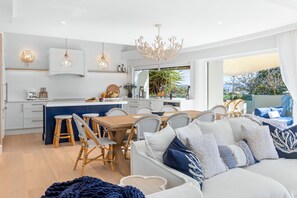 A modern kitchen with white cabinetry, a dining area with wicker chairs, and a large window overlooking a garden.