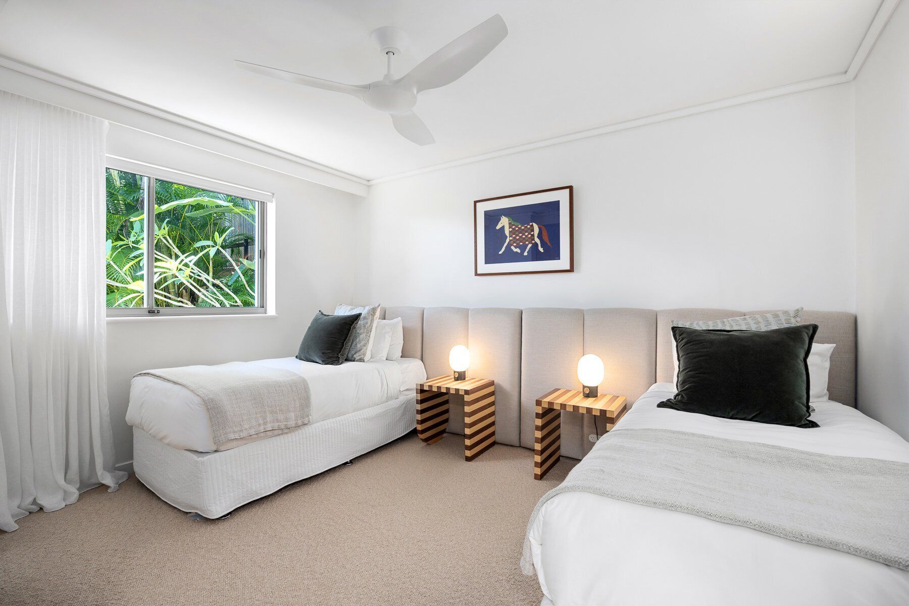 A hotel room with two beds, a ceiling fan, a window with curtains, a framed picture on the wall, and bedside lamps.
