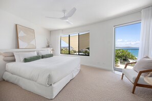 A bedroom with a large bed, a ceiling fan, a view of the sea through a sliding door, and a chair by the window.