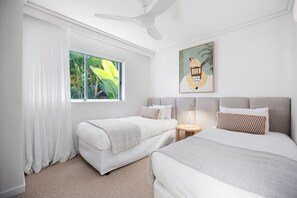 Quarto de hotel com duas camas, ventoinha no teto, janela com cortinas, abajur e um quadro na parede.