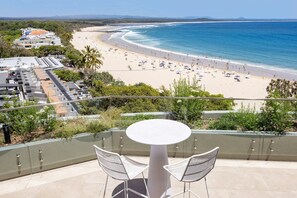 Varanda com mesa e cadeiras voltada para uma praia, com pessoas e um edifício ao fundo.