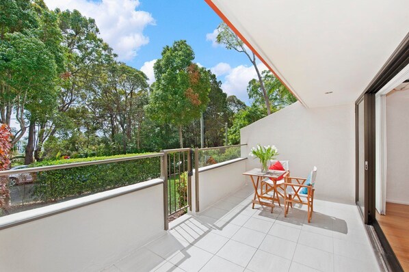 A balcony with a view of a garden and trees, a table with chairs, and a glass railing.