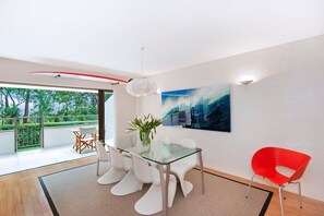 A modern dining area with a glass table, white chairs, a red chair, and a painting on the wall.