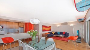 Een moderne woonruimte met een glazen eettafel, witte stoelen, een zithoek en een keuken.