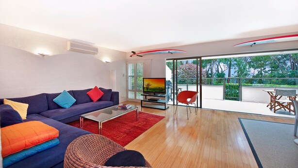 Een moderne woonkamer met een bank, een salontafel en een televisie. Er is een balkon met uitzicht op bomen en een rode stoel.