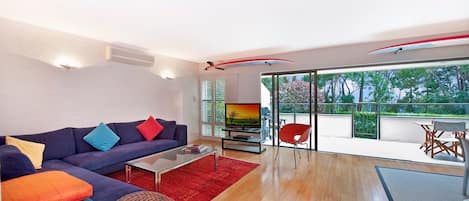 Una sala de estar moderna con sofá, mesa de centro y televisor. Hay un balcón con vista a árboles y una silla roja.