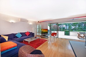 A modern living room with a sofa, a coffee table, and a television. There is a balcony with a view of trees and a red chair.