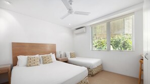Une chambre d’hôtel avec deux lits, un ventilateur de plafond, un appareil de climatisation, une fenêtre avec des stores et une chaise en bois.