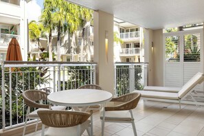 A balcony with a round table and wicker chairs, surrounded by white railings and a view of a building with balconies.