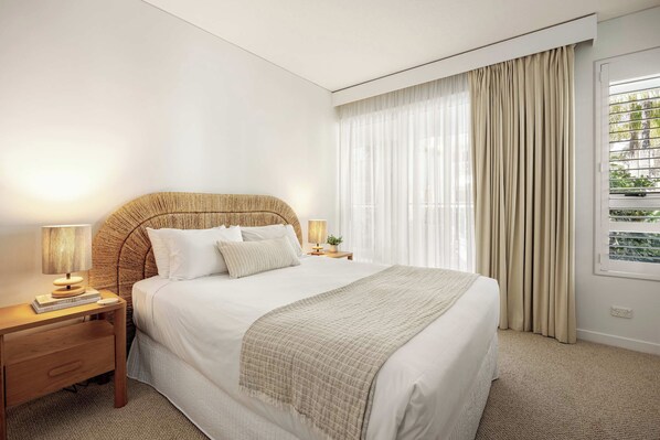 A hotel room with a large bed, a bedside table with a lamp, a window with curtains, and a view of greenery outside.