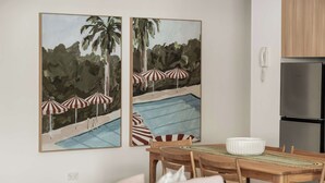 A dining area with a wooden table, chairs, and a white bowl. There are two framed paintings on the wall depicting a pool with umbrellas. A refrigerator is visible in the background. - Laguna 117 - Poolside, Near Beach (Noosa Heads)