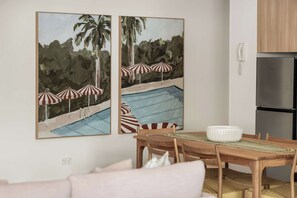 A dining area with a wooden table, chairs, and a white bowl. There are two framed paintings on the wall depicting a pool with umbrellas. A refrigerator is visible in the background.