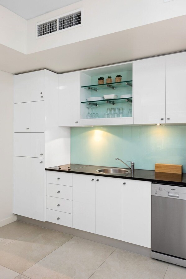 A modern kitchen with white cabinets, a black countertop, a built-in dishwasher, and a glass shelf with kitchenware.