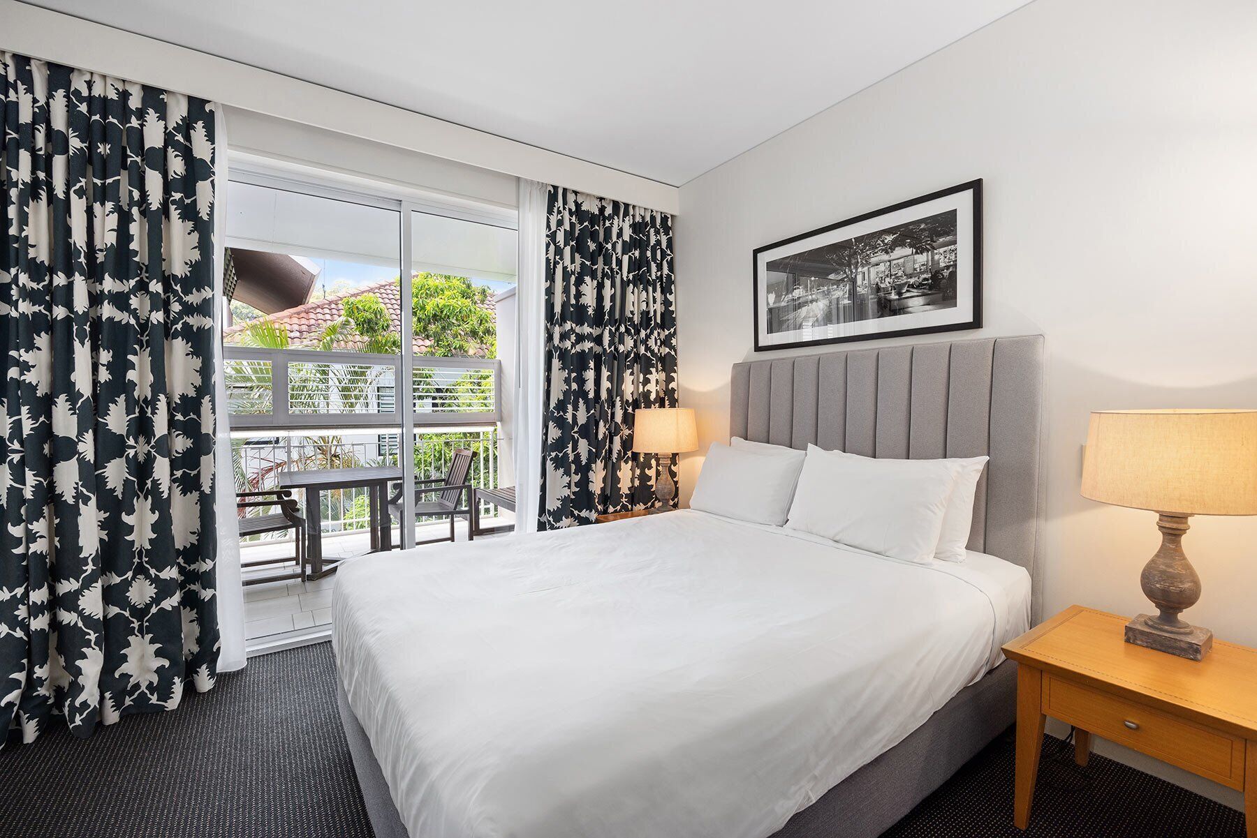 Ein Hotelzimmer mit einem Bett, zwei Nachttischlampen, einem gerahmten Bild an der Wand und einem Balkon mit Tisch und Stühlen.