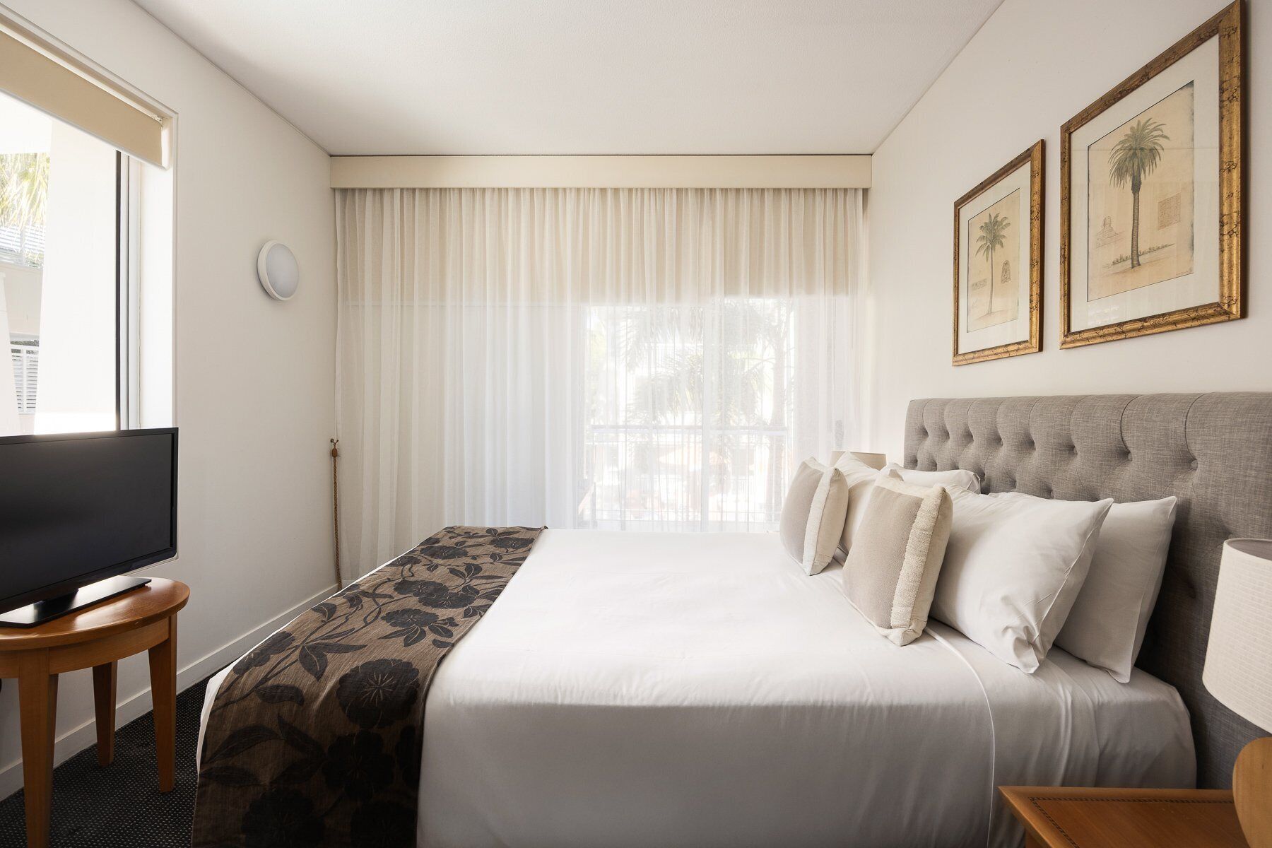 A hotel room with a bed, a television on a wooden table, a window with curtains, and two framed pictures on the wall.