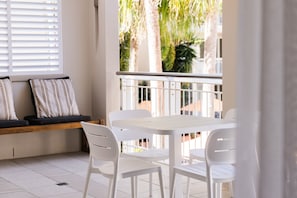 Un balcon avec des chaises blanches et une table, un banc avec des coussins, et une vue sur des palmiers.